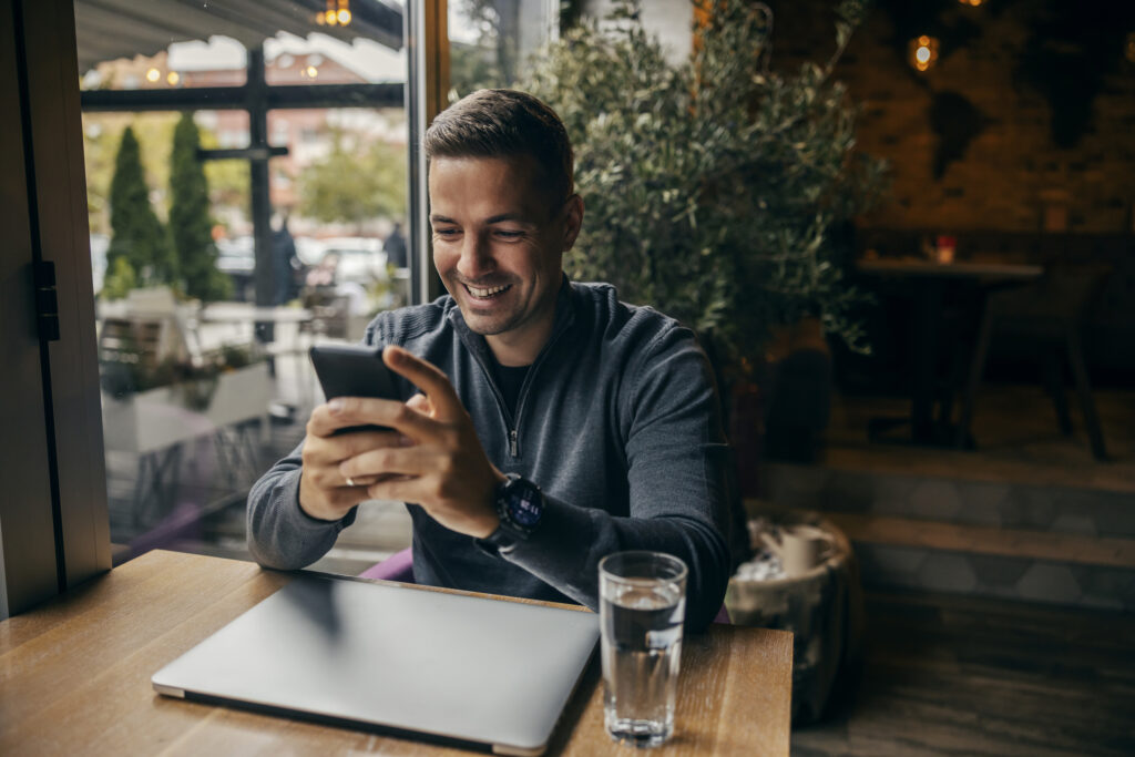 A guy is sitting in cafe and smiling at his phone.